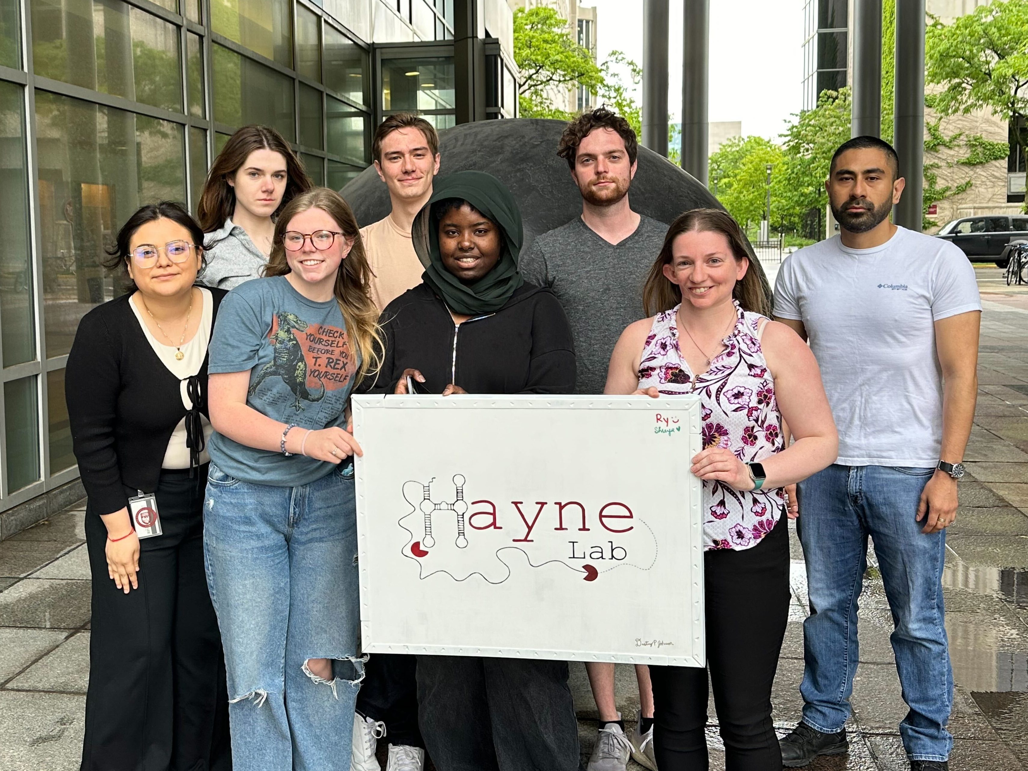 A group photo of the Hayne Lab with a "Hayne Lab" sign being help in front of a large black sphere (art).